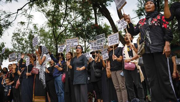 Indische Frauen in teils traditioneller Kleidung protestieren unter freiem Himmel, halten Schilder in der Hand, hinter ihnen Bäume.