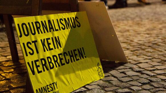 Das Foto zeigt ein Pappschild mit Amnesty-Logo und dem Schriftzug "Journalismus ist kein Verbrechen" auf dem Kopftseinpflaster eines Platzes stehend.