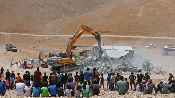 Menschen beobachten von einem Hügel aus, wie ein Bagger ein Haus zerstört.