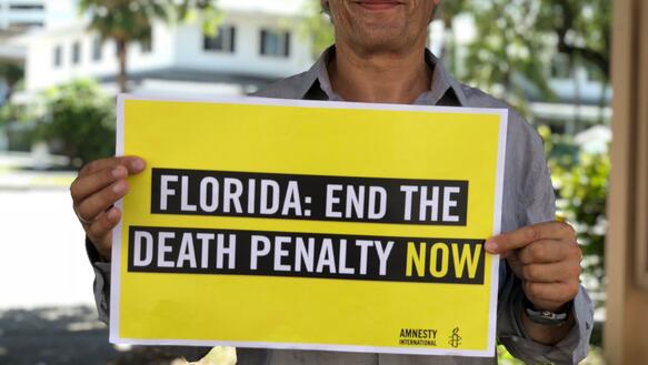 Aufnahme eines Mannes an einem sonnigen Tag. Er blickt leicht lächelnd in die Kamera und hält ein Schild mit Amnesty-Logo vor sich. Auf dem Schild steht: "Florida: End the Death Penalty now"