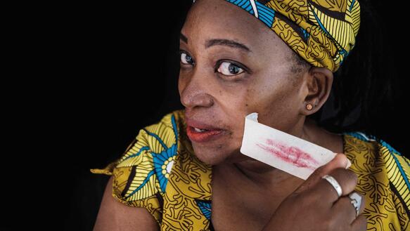 Eine Frau aus Uganda trägt traditionelles Gewand und passendes Kopftuch, sie zieht sich ein Stück Klebeband vom Mund ab, das auf der klebrigen Seite mit Lippenstift verschmiert ist.
