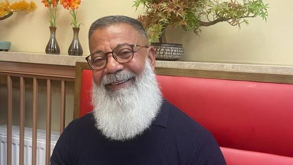 Porträtfoto von Hassan El Sayed, der auf gepolsterten Bank an einem Tisch sitzt. Er hat einen langen Vollbart und trägt Brille und T-Shirt und lächelt in die Kamera.