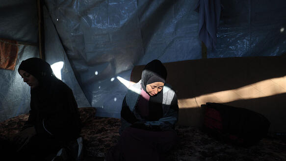 Zwei Frauen mit Kopftüchern sitzen traurig und niedergeschlagen im Halbdunkel in einem Zelt. Sie blicken auf den Boden. Auf das Gesicht der Frau rechts fällt ein Lichtstrahl