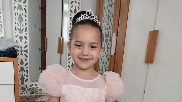 Hind Rajab steht in einem Zimmer. Sie trägt ein Kleid und lächelt in die Kamera. Hinter ihr steht ein Schraank und ein großer Spiegel.k