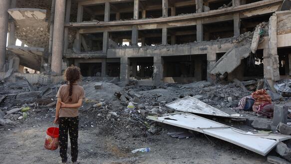 Das Foto zeigt ein von hinten fotografiertes Mädchen, das die Arme hinter dem Rücken verschränkt hat und einen Eimer in der Hand hält. Das Mädchen steht vor der Ruine eines mehrstöckigen zerstörten Gebäudes. Überall liegen Trümmer.