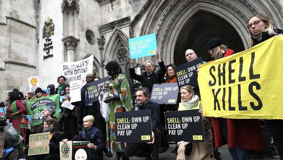 Circa 15 Personen stehen und hocken dich nebeneinander vor einem Gebäuden. Sie halten Schilder und Plakate hoch, auf denen unter anderem steht "Justice for the Niger Delta" oder "Shell kills".