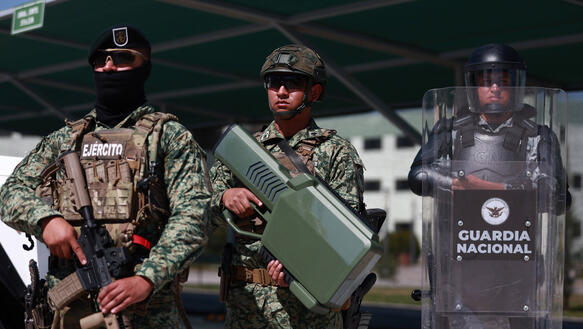 Drei schwer ausgerüstete Mitglieder der mexikanischen Armee und Nationalgarde stehen mit Waffen, taktischem Gerät und einem Schutzschild bei einer Übung nebeneinander.