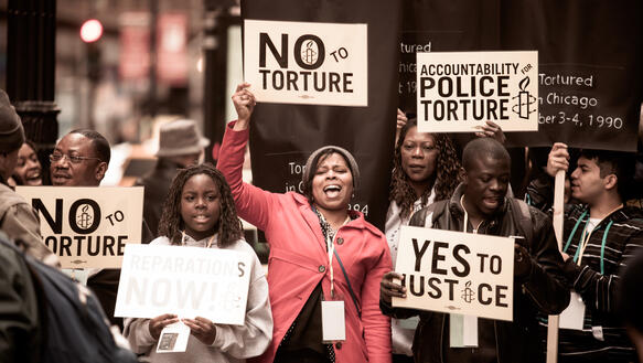 Eine Gruppe von Menschen auf einer Amnesty-Demonstration hält Protestschilder mit Aufschriften wie "No to Torture", "Yes to Justice" und "Accountability for Police Torture"