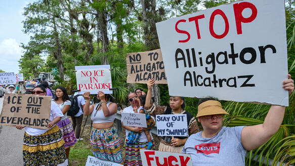 Eine Gruppe von Demonstranten hält am Rande eines Naturgebiets Plakate mit Parolen wie "STOP Alligator Alcatraz" und "ICE OUT OF THE EVERGLADES" in die Kamera. Sie protestieren damit sichtbar gegen die US-Einwanderungsbehörde ICE und ein geplantes Bauprojekt.