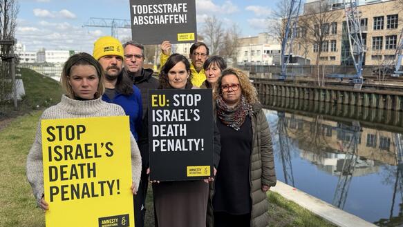 Das Foto zeigt mehrere Personen eng beiannder stehen neben einem Kanal. Im Hintergrund steht ein Kran. Sie halten Schilder mit Amnesty-Logos in die Kamera, auf denen steht: "Stop Israel's death Penalty" und "Todesstrafe abschaffen".