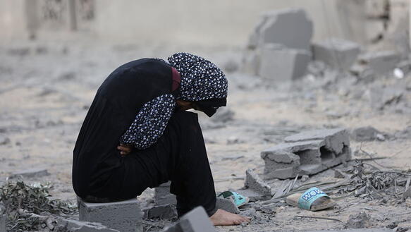 Eine Frau mit Kopftuch sitzt zusammengesunken vor Trümmern.
