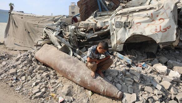 Ein Junge hockt inmitten zerstörter Gebäude auf den Resten einer Rakete.