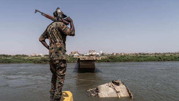 Ein Soldat in Tarnuniform steht auf einem Betonklatz an einem Flussufer. Er hat sein Stumgewehr auf seine rechte Schulter gelegt. Die Brücke vor ihm ist zerstört.
