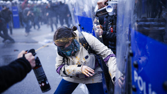 Auf eine vermummte Demonstrantin wird Pfefferspray gespritzt, im Hintergrund Polizei und weitere Demonstrant*innen.
