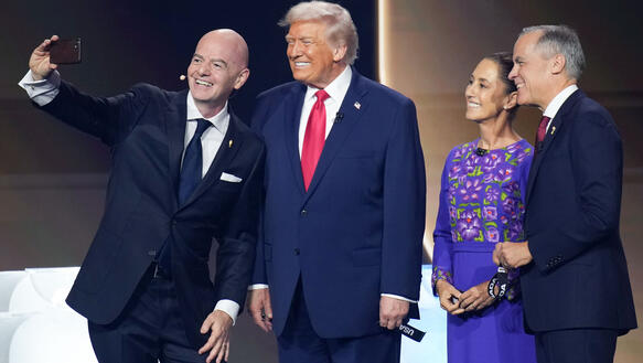 Gianni Infantino nimmt auf einer Bühne ein Selfie mit Donald Trump auf. Sie lächelnd. Neben ihn steht noch eine Frau und ein Mann, die ebenfalls in die Handykamera lächeln.