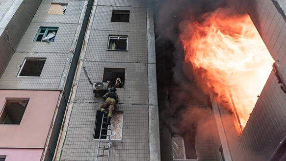 Flammen schlagen aus den Fenstern eines Hochhauses, während Feuerwehrmann auf einer Leiter steht.