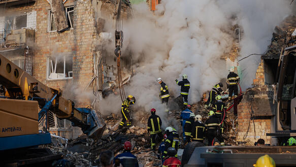 Feuerwehrleute in den Trümmern eines zerstörten Hauses, Rauch steigt auf.