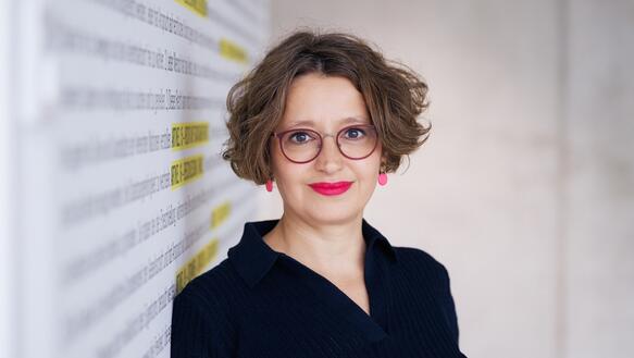 Porträtfoto von Wiebke Gülcibuk, die an eine Wand gelehnt in die Kamera lächelt. Sie trägt eine Brille.
