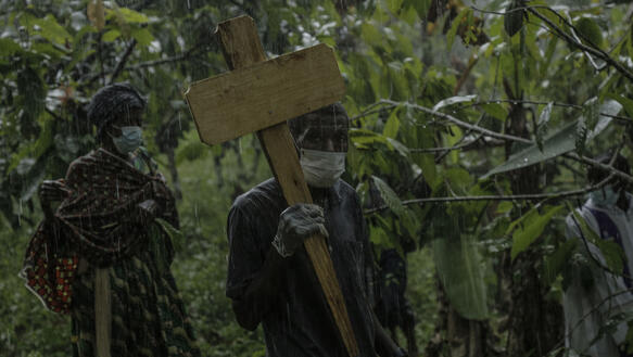 Das Foto zeigt einen Mann mit Mundschutz bei strömenden Regen in einem Dschungel. Er hält ein Holzkreuz in der Hand und schaut in die Kamera.