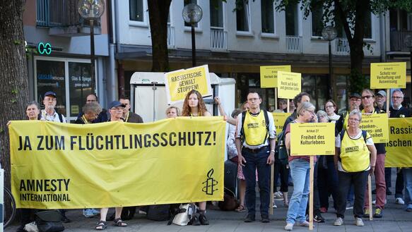 Foto einer Kundgebung mit Personen die ein Banner halten, auf dem steht: "Ja zum Flüchtlingsschutz".