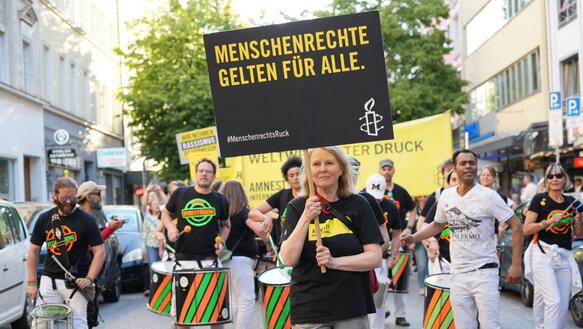 Das Foto zeigt eine Frau an der Spitze eines Demonstrationszuges. Sie hält ein Schild hoch mit Amnesty-Logo und dem Slogan "Menschenrechte gelten für alle!". Hinter ihr spielt eine Trommelgruppe.