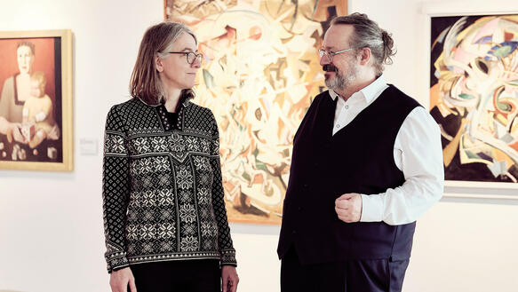 Eine Frau und ein Mann, beide tragen eine Brille, mittleres Alter, stehen in einem Museum, an der Wand hängen gerahmte Bilder und Leinwände.