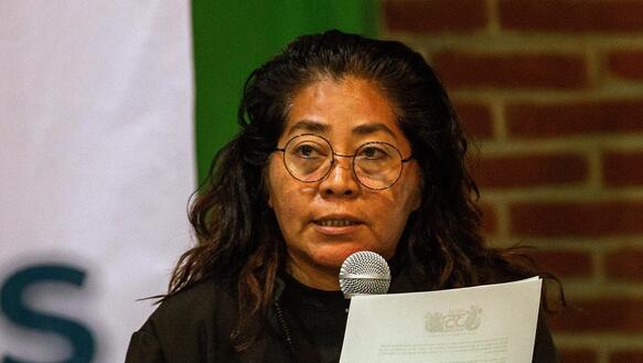 Das Foto zeigt Dora Roblero an einem Tisch sitzend. Sie spricht in ein Mikrofon und hält in der linken Hand einen Zettel. Sie trägt eine Brille.