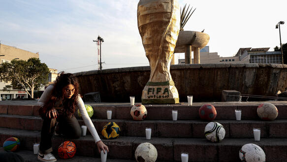 Kunstaktion in Mexiko: Eine Studentin stellt eine Kerze zu einer großen FIFA-WM-Pokal-Nachbildung, die an vermisste Personen erinnert und von Fußbällen umgeben ist.
