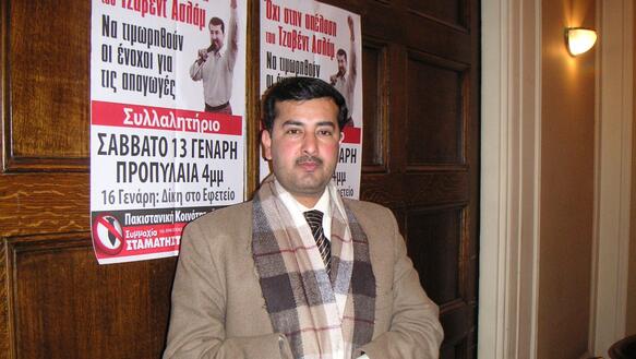 Foto von Javed Aslam, der Jacke, Schal und Krawatte trägt und in seinen gefalteten Händen ein Blatt Papier vor sich hält. Er steht vor einer holzvertäfelten Wand, auf der zwei Poster angebracht sind mit einem Mann, der seine linke Faust in die Höhe streckt.