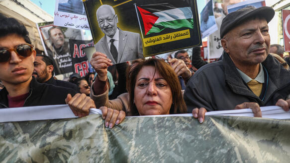 Das Foto zeigt einen jüngeren Mann, eine Frau und einen Mann, die bei einer Demonstration ein Banner vor sich hertragen. Hinter ihnen hält eine Person ein Plakt hoch mit dem Fotos Mannes.