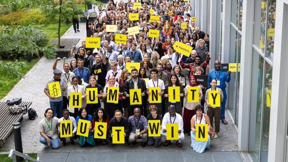 Gruppenfoto mit zig Menschen neben einem Gebäude. Einige halten Schilder hoch. Die Personen vorne halten Tafeln mit Buchstaben hoch, die die Slogan bilden: "Humanity must win."