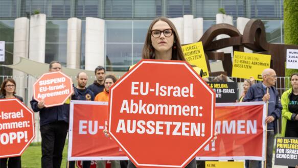 Menschen bei Protestaktion vor dem Kanzleramt in Berlin mit Transparenten und Schildern. Eine Frau steht im Vordergrund. Auf ihrem Schild steht: "EU-Israel-Abkommen aussetzen!"