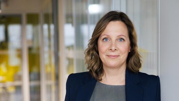 Porträtfoto von Julia Duchrow, aufgenommen in einem lange  Gang in einem Bürogebäude. Sie lehnt mit ihrem rechten Arm an eine Wand und lächelt in die Kamera-