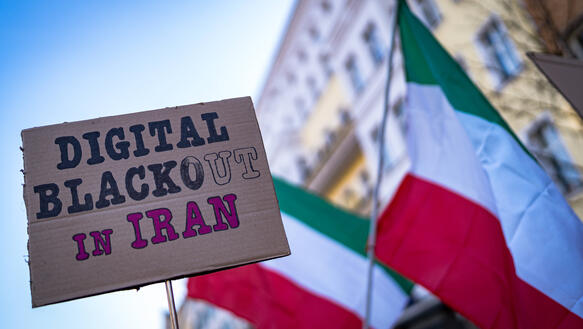 Eine Nahaufnahme eines Pappschildes mit der handgeschriebenen Aufschrift „DIGITAL BLACKOUT IN IRAN“, gehalten vor einem unscharfen Hintergrund mit iranischen Flaggen und der Fassade eines europäischen Gebäudes während einer Demonstration.