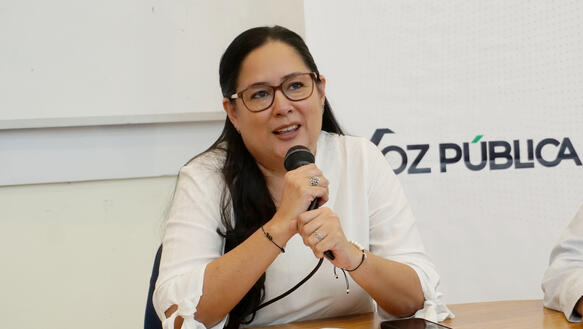 Das Foto zeigt Ruth López an einem Tisch sitzend. Sie spricht in ein Mikrofon, das sie mit beiden Händen vor sich hält. Sie trägt eine Brille und lächelt leicht.