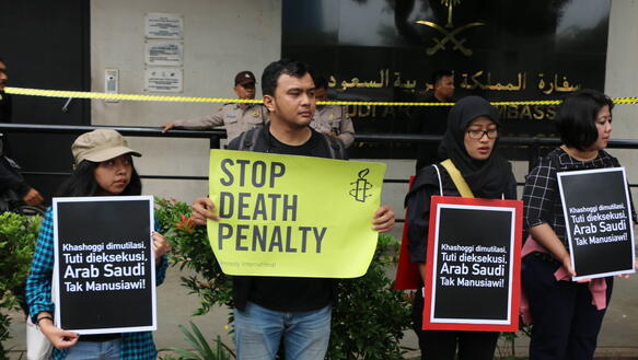 Vier Demonstrierende stehen nebeneinander vor dem Gebäude der saudi-arabischen Botschaft in Jakarta und halten Protestschilder in die Kamera. Eine Person in der Mitte hält ein großes, leuchtend gelbes Schild mit der Aufschrift „STOP DEATH PENALTY“ und dem Logo von Amnesty International.