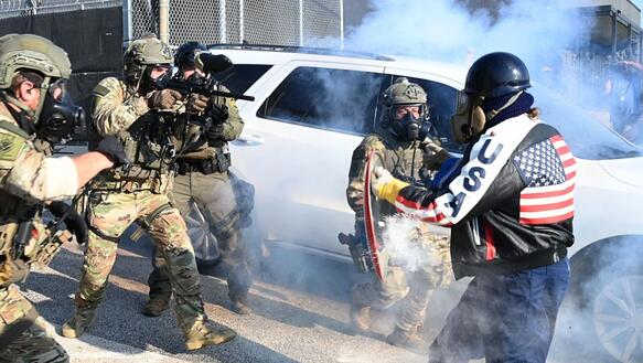 Bewaffnete Polizeikräfte gehen auf eine Person mit einer Gasmaske, einem Schild und einer Motorradjacke auf der "USA" steht zu.