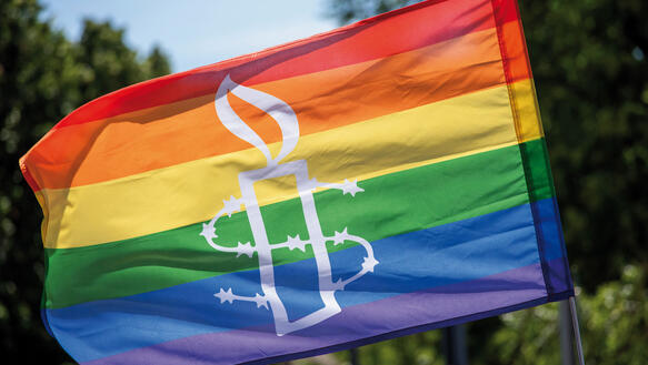 Eine Regenbogenfahne in der Sonne, darauf das Symbol von Amnesty International, eine stacheldrahtumwundene Kerze.