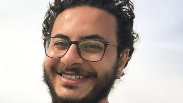 Porträt eines Mannes mit dunklen Locken und Brille, der in die Kamera lächelt.