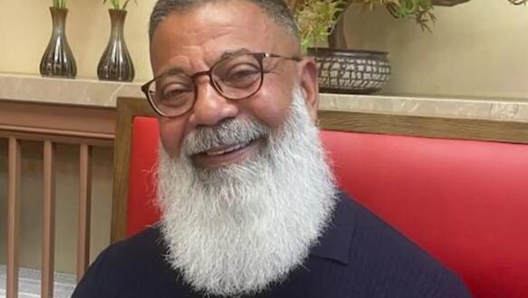 Porträtfoto von Hassan El Sayed, der auf gepolsterten Bank an einem Tisch sitzt. Er hat einen langen Vollbart und trägt Brille und T-Shirt und lächelt in die Kamera.