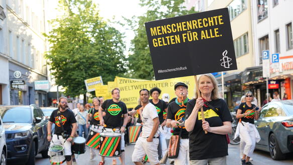 Mehrere Menschen mit gelben Protestschilder, dazu Percussion-Band, eine Frau im Vordergrund mit dem Schild "Menschenrechte gelten für alle"