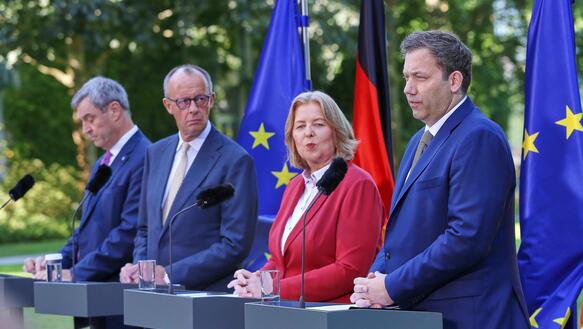Markus Söder, Friedrich Merz, Bärbel Bas und Lars Klingbeil stehen nebeneinander im Freien vor kleine Podien mit Mikros. Hinter ihnen steht eine Deutschland-Flagge und zwei Flaggen der Europäischen Union.