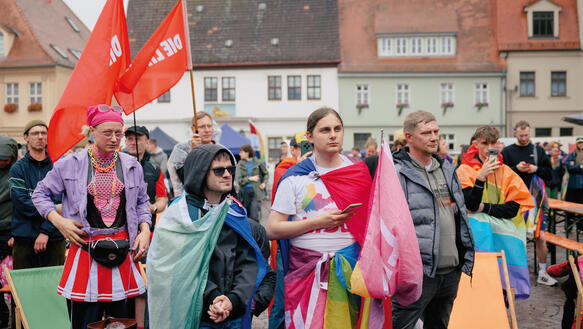 Teilnehmende des CSD stehen auf einem Marktplatz in der Provinz, der Boden regennaß, sie halten teilweise Fahnen mit "Die Linke" beschrieben.