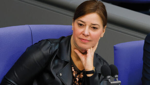 Eine Frau in Lederjacke sitzt auf einem Stuhl im Deutschen Bundestag und stützt ihr Kinn auf der linken Hand ab, vor ihr zwei Tischmikrofone.