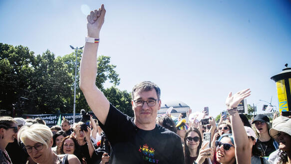 Ein mittelalter Mann reckt seinen Arm in die Höhe, er steht mitten in einem Protestzug zwischen Menschen, es ist Sommer, unter freiem Himmel, im Hintergrund Bäume.