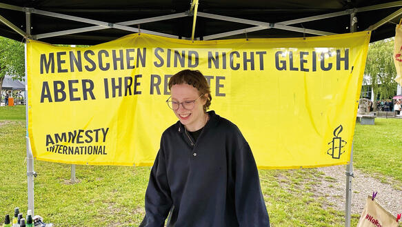 Eine junge Frau mit Brille steht unter dem Pavillon eines Amnesty-Aktionsstands und lächelt, auf einem Banner hinter ihr steht: Menschen sind nicht gleich, aber ihre Rechte