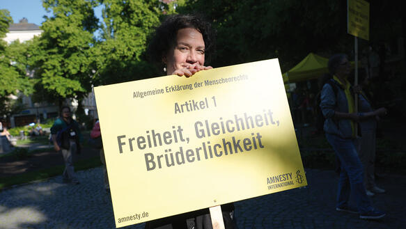 Eine Person steht hinter einem Demo-Schild von Amnesty, auf dem steht: "Freiheit, Gleichheit, Brüderlichkeit"