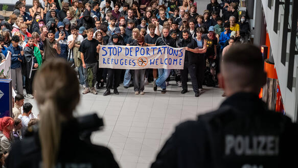 Menschenmenge mit Protestbanner, im Vordergrund zwei Polizeibeamt*innen