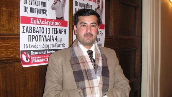 Foto von Javed Aslam, der Jacke, Schal und Krawatte trägt und in seinen gefalteten Händen ein Blatt Papier vor sich hält. Er steht vor einer holzvertäfelten Wand, auf der zwei Poster angebracht sind mit einem Mann, der seine linke Faust in die Höhe streckt.