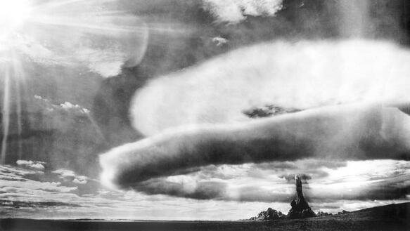 Der Wolkenring einer Atombomendetonation breitet sich über einer flachen Landschaft aus, im Hintergrund steigt ein Rauchpilz auf.